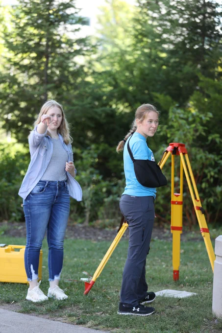 Two students work outdoors with surveying equipment on a grassy area. One points ahead while the other stands near a yellow tripod, wearing a sling, as they prepare to take measurements among trees.
