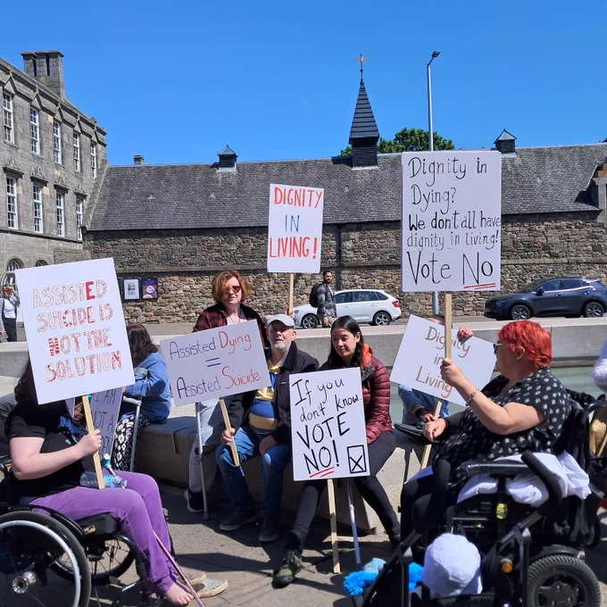 GDA  members are gathered at the Scottish Parliament to protest the Assisted Dying Bill in May 2025. It's a bright and sunny picture, and members hold placards that read: "Dignity in Dying? We don't all have dignity in Living! Vote No", "If you don't know vote no! "Dignity in Living" and "Assisted suicide is not the solution"