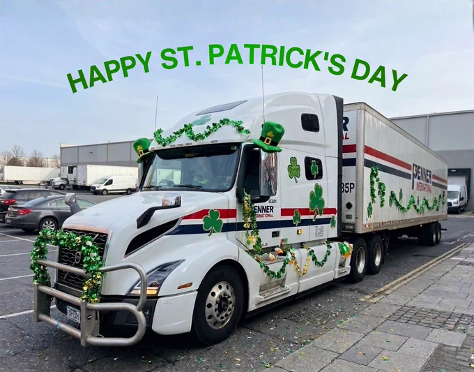 A white Penner International transport truck is parked in a lot and decorated for St. Patrick’s Day. The truck is covered in green garlands, shamrock cutouts, small green hats, and festive tinsel. The trailer behind it is also wrapped with green garland. Above the truck in the image, large green text reads “HAPPY ST. PATRICK’S DAY.