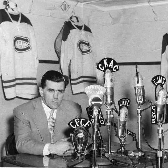 Maurice Richard sits at a table during a press conference, leaning forward with his arms crossed and looking toward the cameras. Several vintage broadcast microphones crowd the table in front of him, each labeled with radio station call signs including CFCF, CKAC, CKVL, CJMS, and Radio Canada. Behind him, three Montreal Canadiens jerseys hang on hooks along the wall, one showing the number 9. The room appears to be a locker room or team area, with bare walls and hooks visible above the jerseys.

