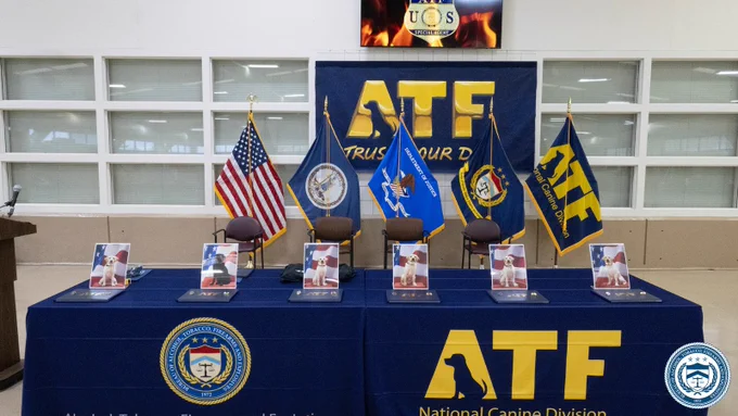 Table set for a National ATF K-9 Graduation event with flags, chairs, and framed photos of graduates on a blue cloth featuring the ATF logo and seal.