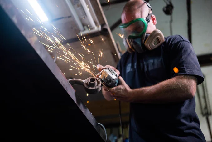 A worker in a protective mask using an angle grinder, emitting sparks in a workshop setting.