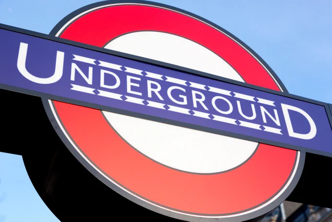 A London Underground sign. It features a red circle bisected by a horizontal blue bar with the word \"UNDERGROUND\" in white capital letters. The blue bar has a repeating pattern of white diamonds running along the top and bottom edges.
