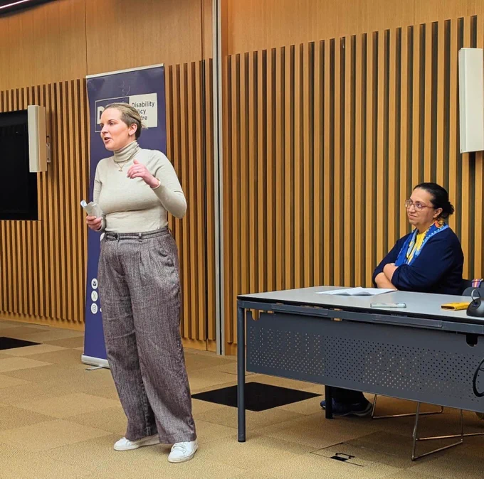 Emma stands speaking at a City Hall roundtable. She has blonde hair and wears a beige jumper, holding notes. Hina, wearing blue, sits beside her listening. A Disability Policy Centre banner stands behind.