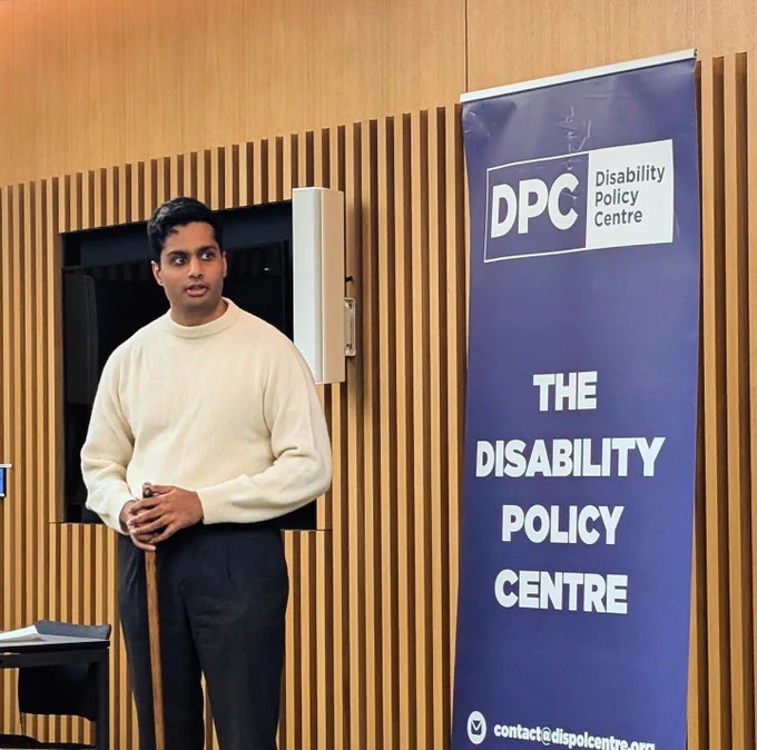 Arun stands beside a Disability Policy Centre banner at City Hall, wearing a white jumper, addressing the room during a roundtable discussion in a modern meeting space.