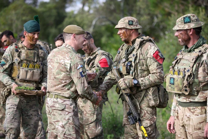 Military personnel in camouflage gear and face paint are gathered outside. Some have red armbands and various equipment. Two individuals in the center are shaking hands. The scene is set in a grassy area with trees in the background.