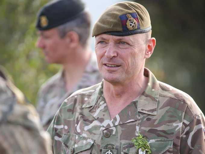 Military personnel in camouflage uniforms are engaged in conversation. One is wearing a Shamrock on his shirt pocket.  The scene is set in a grassy area with trees in the background.