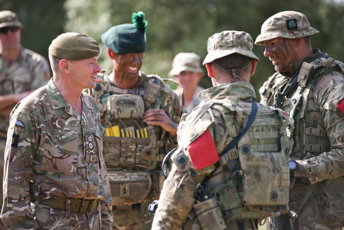 A group of military personnel in camouflage uniforms are standing outside, talking. They are wearing tactical gear, including vests and helmets, and their faces are painted for camouflage.
