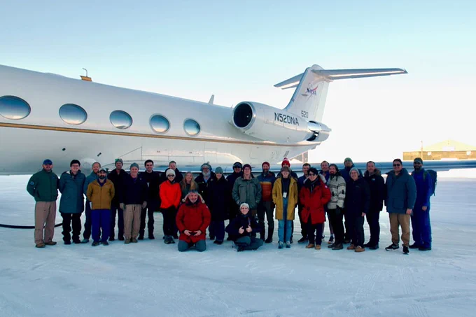 A group of people is standing and kneeling on a snowy surface in front of a NASA aircraft on a clear day. The plane has "N520NA" on its tail. Some individuals are dressed in winter clothing, suggesting a cold environment. A building is visible in the background.