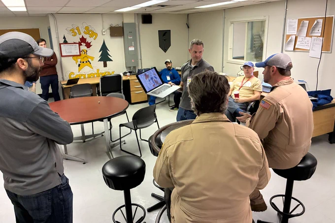 A group of people are gathered in a room. One person is holding a laptop, showing an image to others seated around a table. The room has bulletin boards and a window.