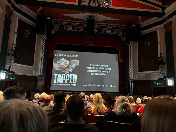 Theatre hall with audience members viewing a screening with the words Tapped on screen
