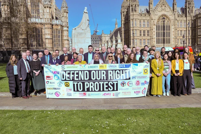Image shows a large group of people outside Westminster standing behind a banner reading \"Defend our right to protest\" 