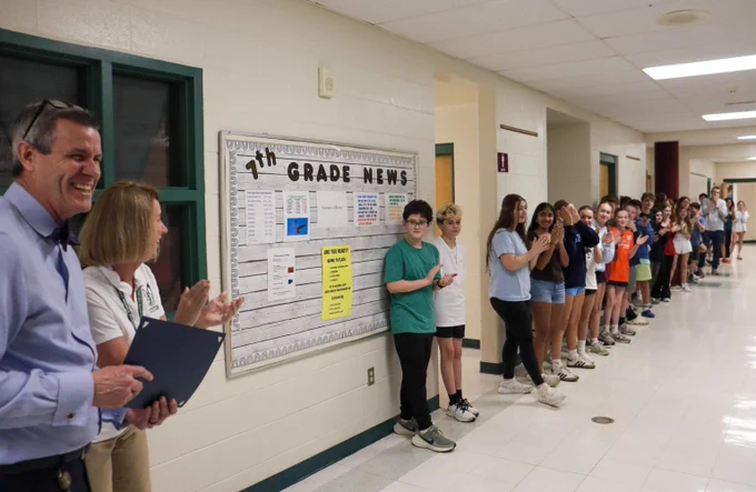 Students and teachers lined up along a school hallway wall near a bulletin board.
