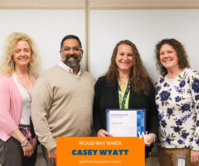 Four colleagues stand together in an office, with one woman holding a certificate of achievement for Casey Wyatt from Spellman Education Center.