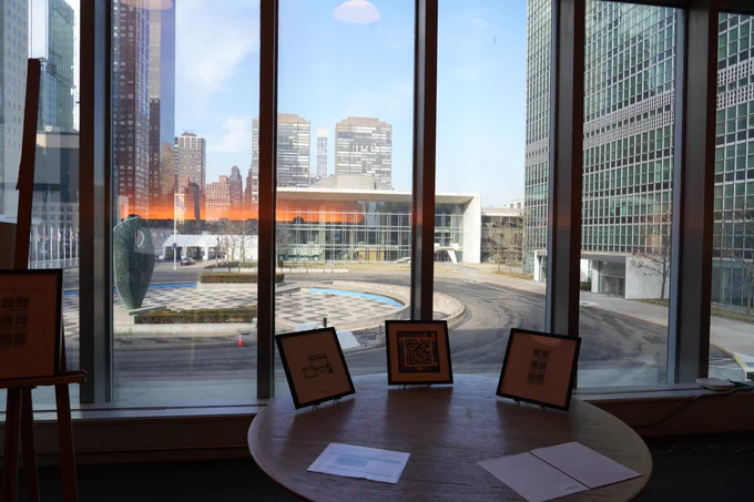 A round wooden table stands in front of tall floor‑to‑ceiling windows overlooking a plaza with modern buildings. Three framed items or printed displays sit on the table, each propped upright. Outside the window, the plaza, sculptures, flags, and surrounding skyscrapers are visible under a clear sky.