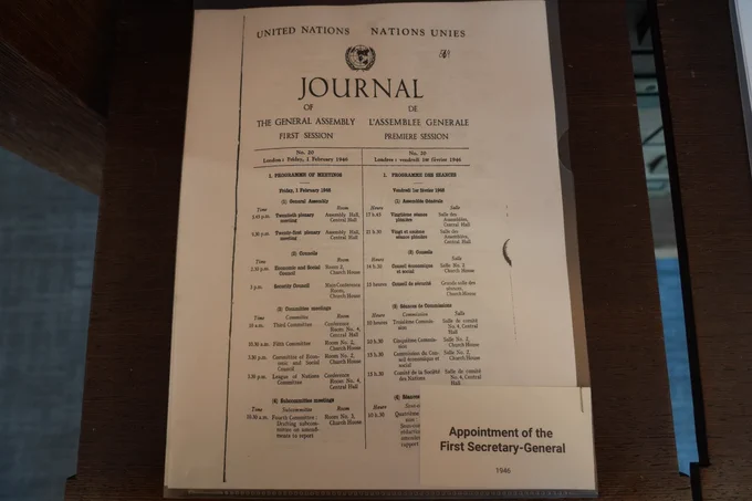 A close‑up view of an archival document titled “Journal of the General Assembly — First Session,” with text in English and French under the United Nations emblem. The document lists meeting schedules and agenda items dated February 1946. A small label beneath the document reads “Appointment of the First Secretary‑General — 1946.”