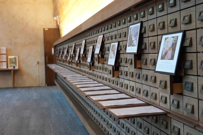 A long wall of wooden card‑catalog drawers in a softly lit room, each drawer with a metal pull handle. Several drawers along the upper row are pulled open and display mounted photographs or documents. A row of flat archival folders is laid out on an extended shelf beneath the drawers. The setting resembles a museum or archival exhibition space.