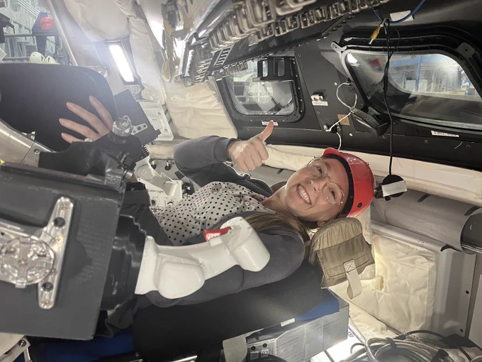 Allison is seated in the Orion spacecraft mockup at the JSC Space Vehicle Mockup Facility. She wears a hard hat and gives a thumbs-up to the camera