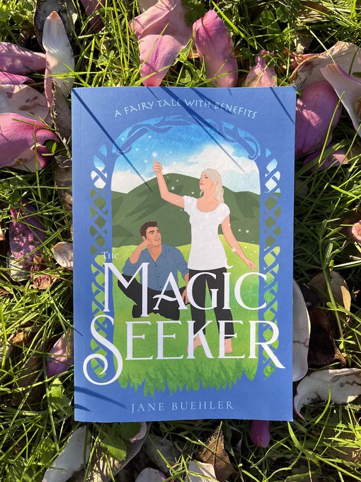 the book The Magic Seeker resting on grass and purple magnolia blossom petals; the book cover shows a drawing of a man sitting on grass and watching a woman who lifts her hand to the sky amid floating sparkles