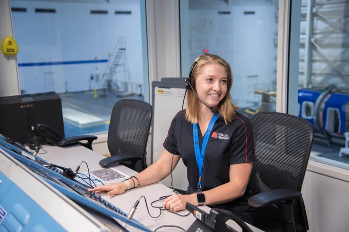 Allison smiles wearing a headset as she sits at a table with controls in front of her. The UNL Air & Space Research Team was at the NASA Neutral Buoyancy Lab to test their design as finalists for the Micro-g Neutral Buoyancy Experiment Design Teams Competition. 