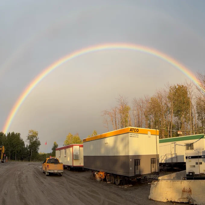 A construction site with several trailers and equipment. Above, a vivid double rainbow arches across a grey sky, with trees lining the background.
