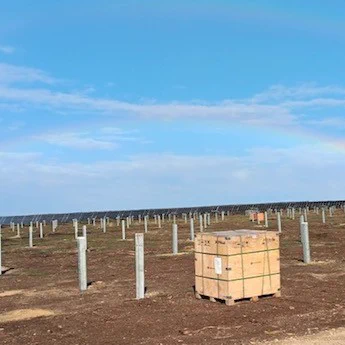 A large field with rows of metal posts, possibly for solar panels, under a wide, clear sky. A faint rainbow arcs across the horizon. In the foreground, there is a wooden crate on the ground.