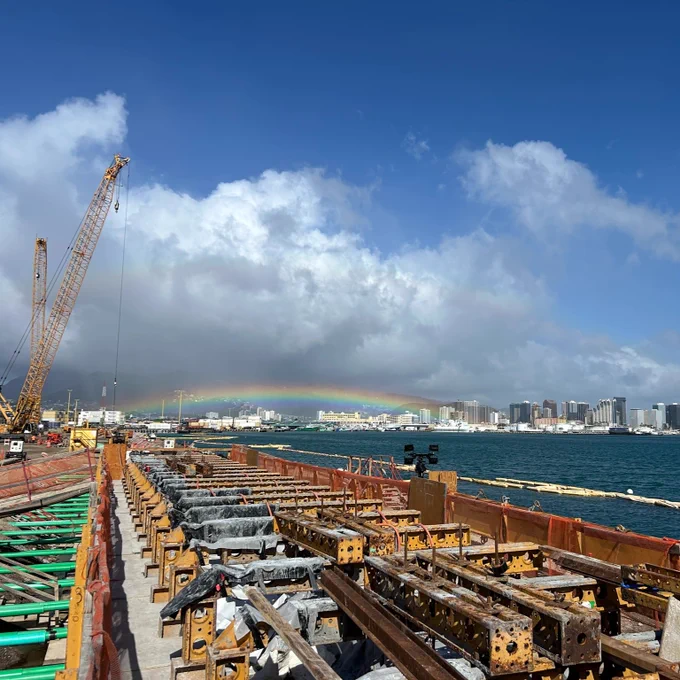 A construction site near a waterfront with cranes and metal structures under a blue sky with clouds. A rainbow is visible over the city skyline in the background.
