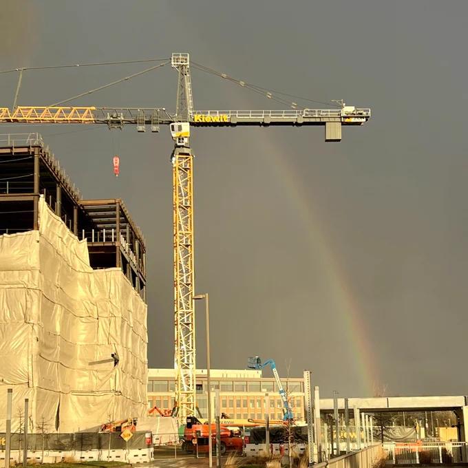 A construction site with a partially built structure, covered in protective material, is highlighted by a rainbow. A large yellow crane with the name "Kroll" is visible. In the background, there's a multi-story building and cloudy sky.