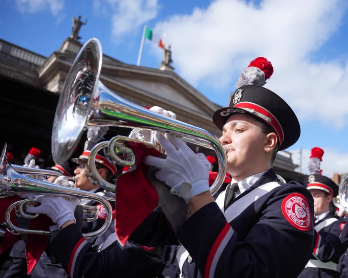 Mellophone member marches by a building with an Ireland flag up top.