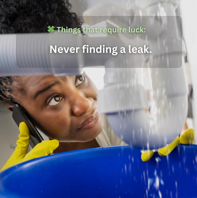 A person wearing yellow gloves observes a leaking pipe beneath a sink, holding a blue bucket to catch water. They are also talking on a phone. A text overlay reads, "Things that require luck: Never finding a leak.