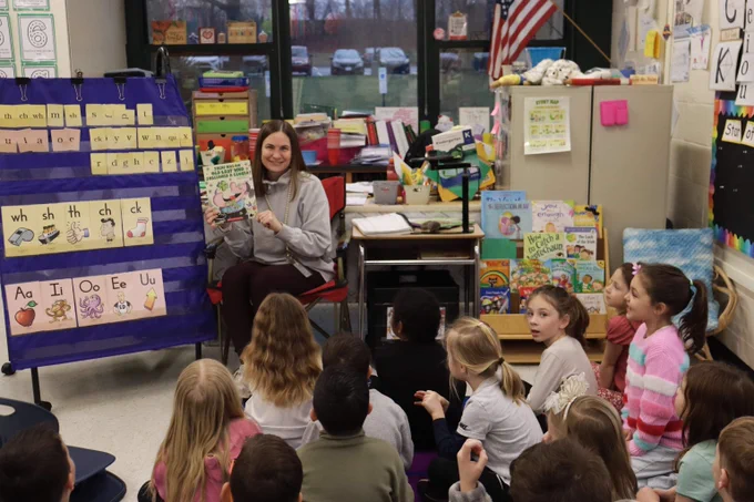 Teacher sitting in front of a classroom reading a book to attentive young students seated on the floor.