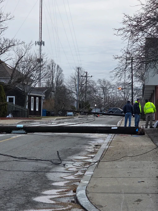 A large utility pole lies across a residential street, pulling down power lines, while several people in high-visibility jackets stand nearby on the sidewalk.