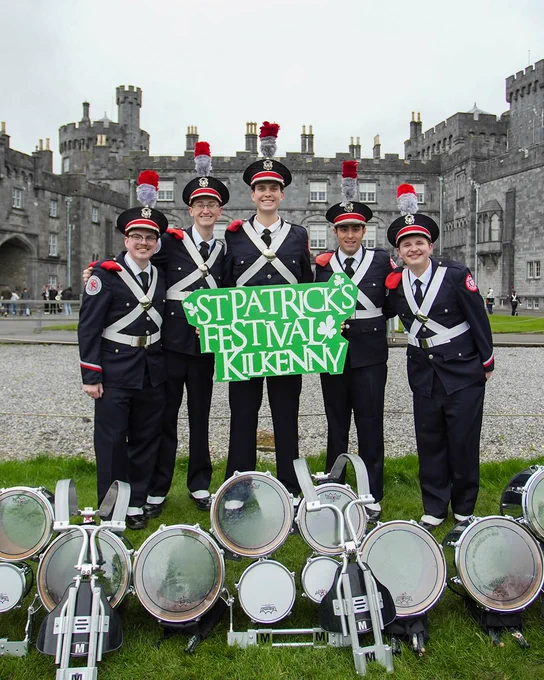 Members of the marching band holding a sign that reads "St. Patrick's Festival Kilkenny." They are standing behind their drums and in front of a castle.