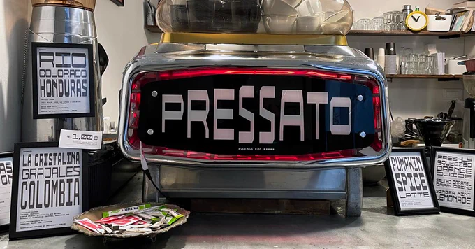 A coffee machine with large white letters reading "PRESSATO" is prominently displayed on a counter. Surrounding the machine are various framed signs. Coffee-making equipment and small decor items are visible in the background.
