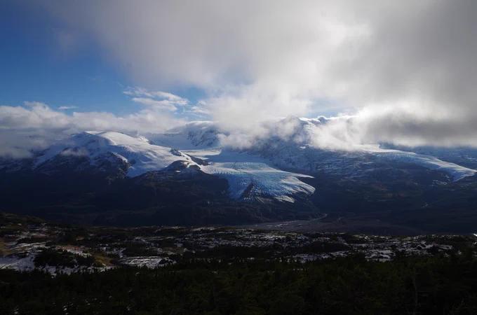 Snow-covered mountain range with a large glacier under a cloudy sky partially revealing blue patches.