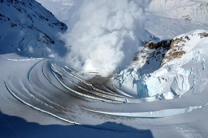 Steam rising from a volcanic crater surrounded by snow-covered mountains and icy terrain.