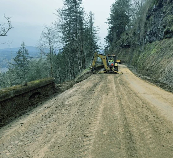 An excavator removes slide debris from the Historic Columbia River Highway pushing the debris from the roadway onto the cliffside below. Tall trees line both sides of the road, with a cliff on the left. Overcast sky above. 