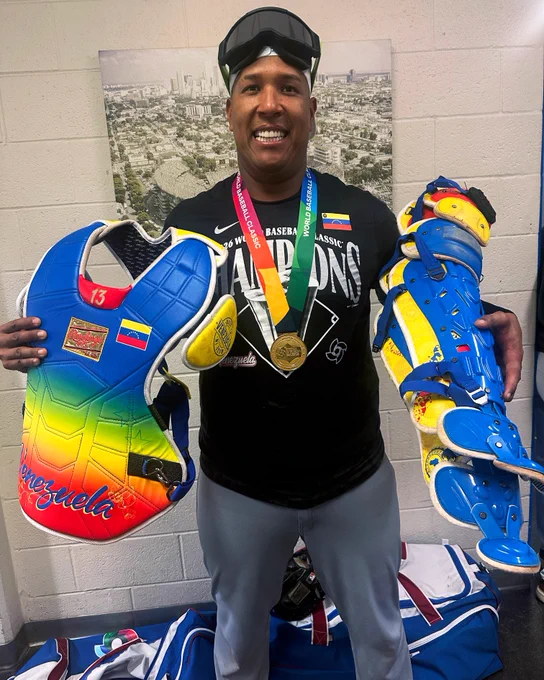 Salvador Perez holds a blue, yellow and red accented chest protector with Venezuela written in blue script and symbolism inspired by the nation’s flag. In his opposite arm, he holds a pair of blue and yellow shin guards.