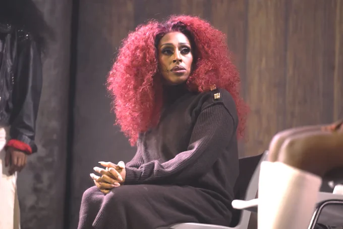 Performer Alexandra Burke sits on stage, dressed as Chaka Khan and wearing a reddish wig.
