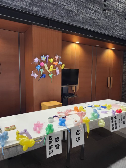 ボランティア交流会・受付の写真です。
白い長机が２つあり、その上に沢山のバルーンアートが置かれています。机奥の柱にも花やプードルなど１５個ほどのバルーンアートが飾られています。