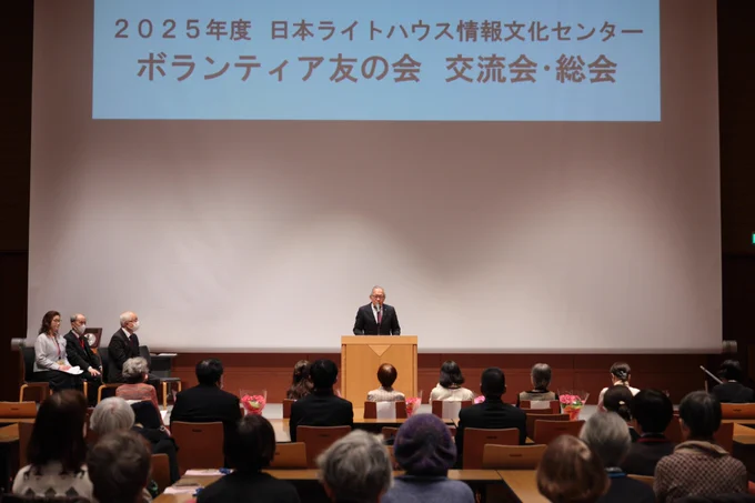 ステージ中央に演台があり、当法人の理事長が挨拶をおこなっています。
ステージ奥のスクリーンには2025年度日本ライトハウス情報文化センターボランティア友の会　交流会・総会と表示されています。
ステージの手前には鉢植えのお花が並んでいます。