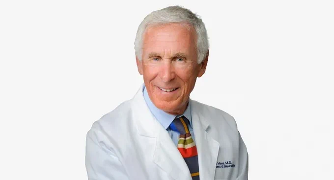 Dr. Brent Basel, clinical professor of neurology at The University of Texas Medical Branch (UTMB), smiles behind a white background. He's wearing his whitecoat and a striped tie.  