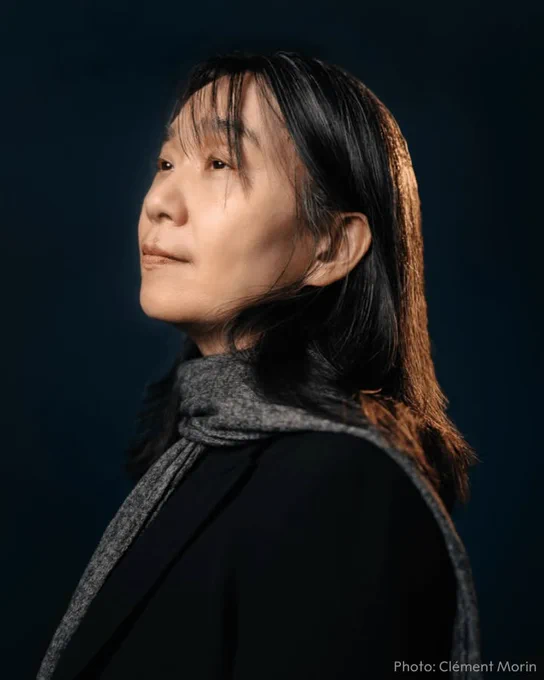 Han Kang in profile, looking to the side. She has shoulder-length black hair and is wearing a dark jacket and grey scarf against a dark background.