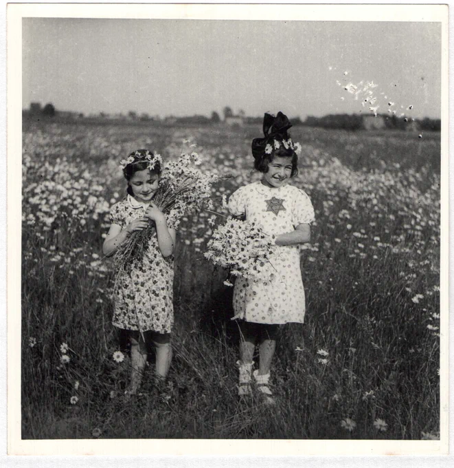 Two young girls holding bouquets of wildflowers in a meadow, wearing dresses and bows in their hair, smiling joyfully.