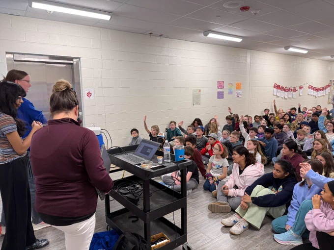 Teacher presenting to a large group of attentive students seated on the floor in a classroom setting.
