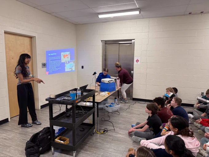 A student stands near a projected presentation while a group of children sit on the floor attentively listening in a classroom.