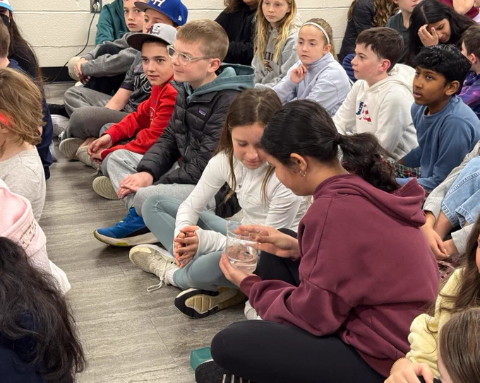 A group of children sitting on the floor in a classroom, attentively watching an activity involving a clear container.