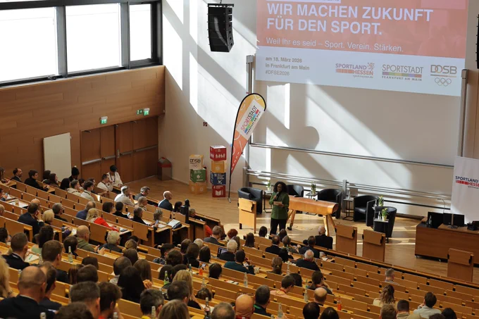 Das Plenum sitzt in einem Hörsaal. Vorne auf der Bühne steht die Moderatorin mit einem Mikrofon. Foto: DOSB