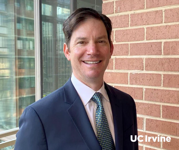 New UC Irvine Physical Sciences dean Seth Cohen is standing in front of a brick wall and glass windows, wearing a suit and tie. The text "UC Irvine" is visible in the corner.