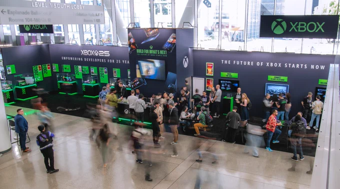 A busy Xbox GDC booth with people walking, playing games, and talking. Large Xbox banners and glowing green displays line the space, highlighting "Xbox 25" and “The Future of Xbox Starts Now.” (Alt generated by Microsoft Copilot and reviewed for accuracy)